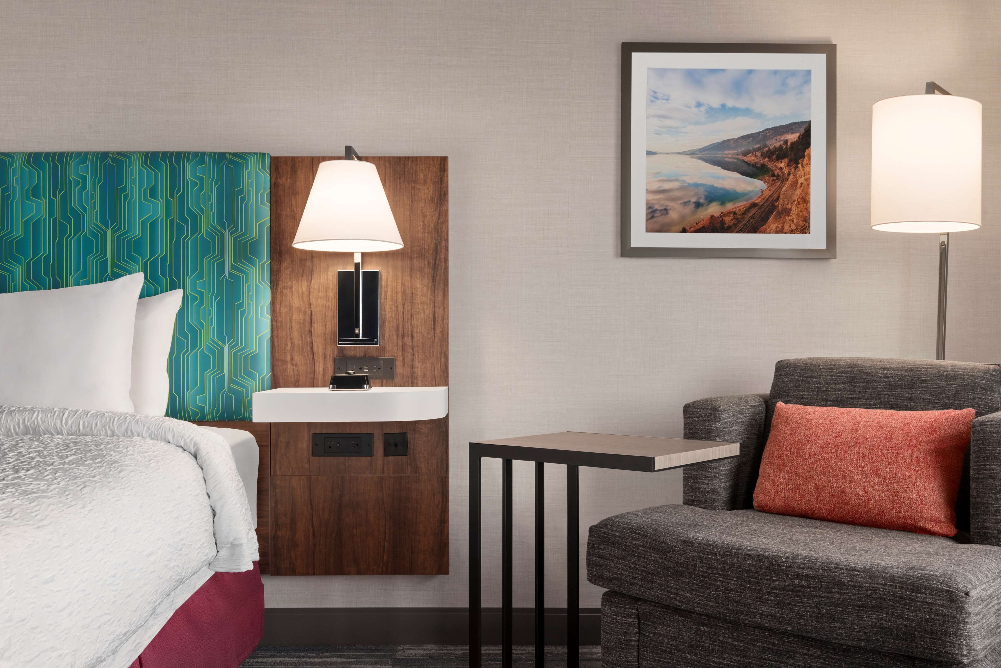 Quarto de hotel com cama, escrivaninha, cadeira, pia fixada na parede e quadro emoldurado.