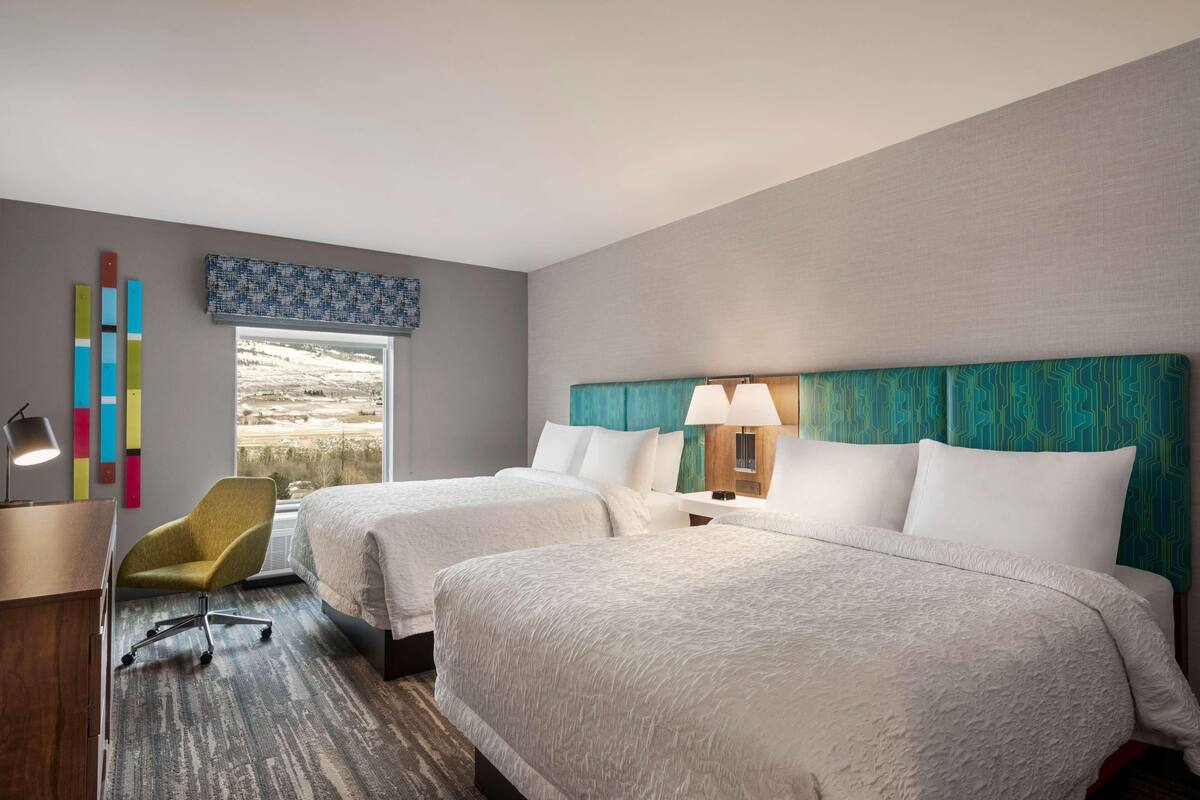 A hotel room with two beds, a desk, a chair, and a view of a landscape.