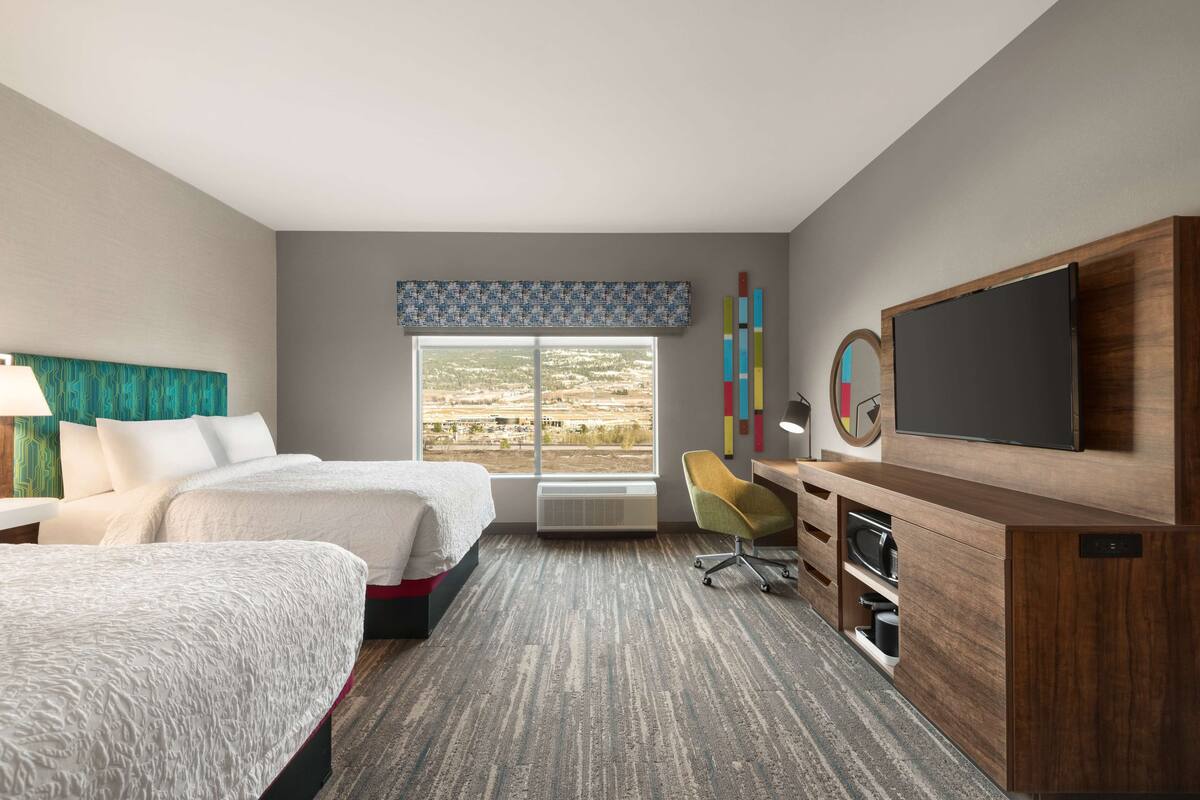 A hotel room with two beds, a desk with a TV, a chair, and a large window with a view of the landscape.