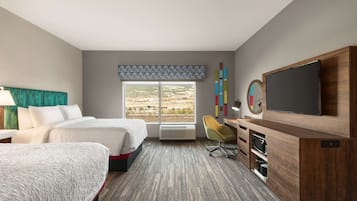 A hotel room with two beds, a desk with a TV, a chair, and a large window with a view of the landscape.