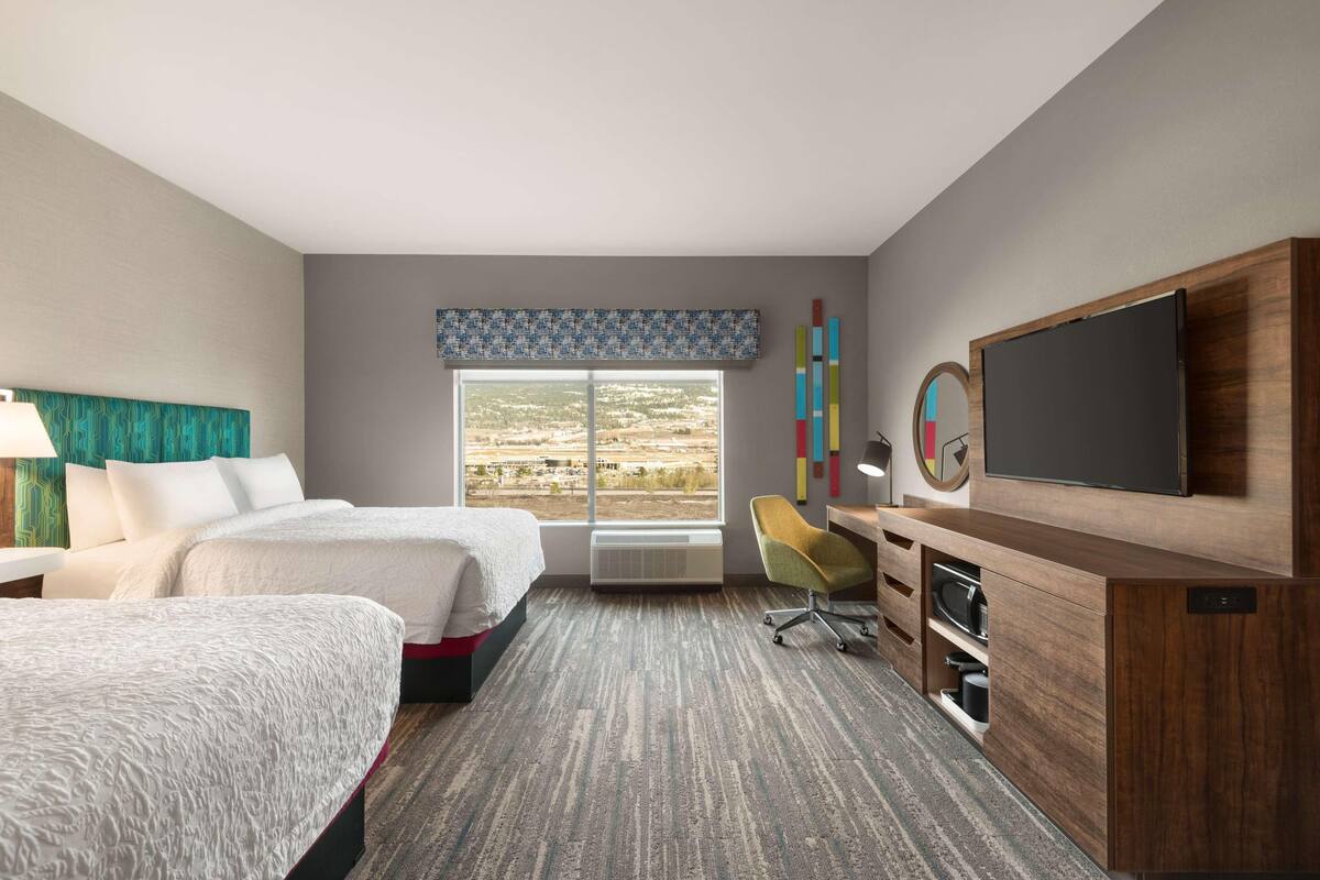 A hotel room with two beds, a desk with a flat-screen TV, a chair, and a large window with a view of the landscape.