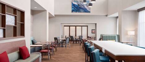 Um saguão de hotel com uma longa mesa de jantar, várias cadeiras, um sofá com almofadas vermelhas e uma grande pintura abstrata na parede.