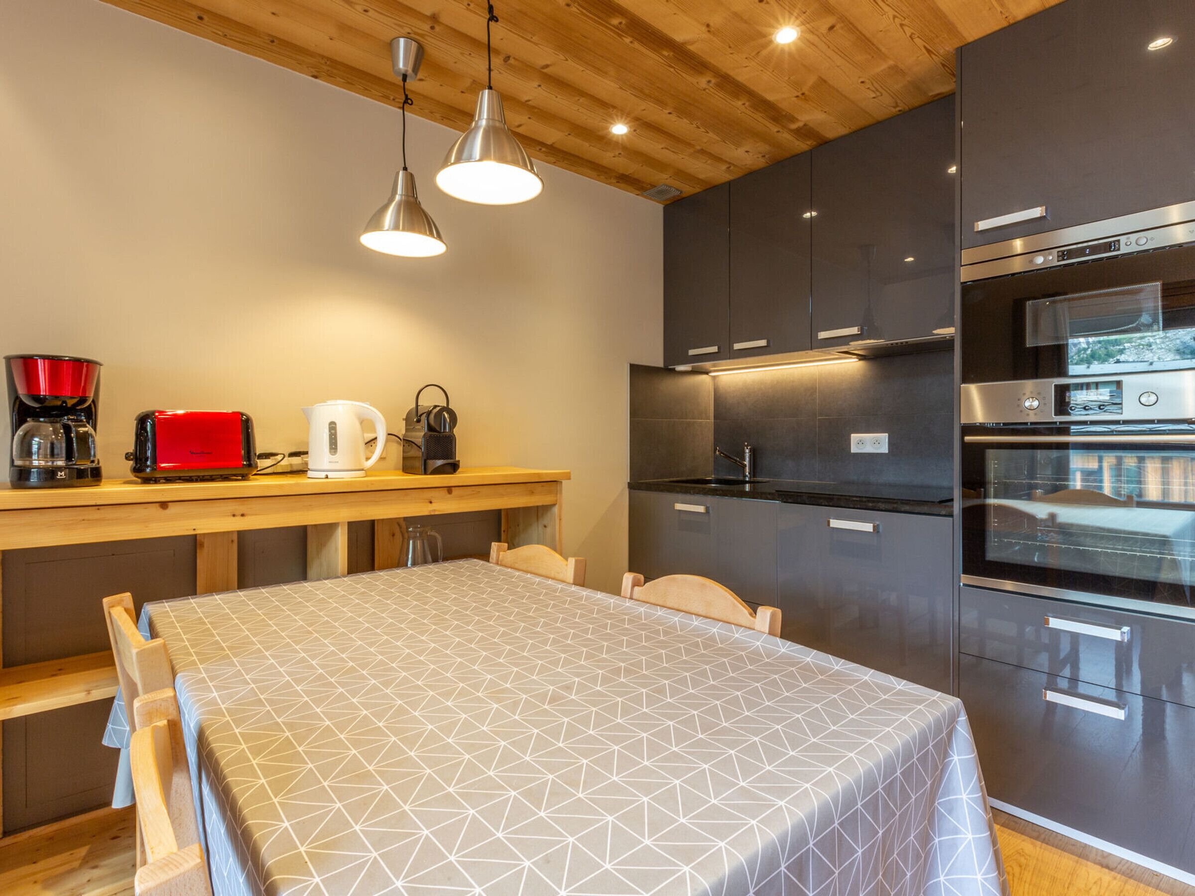 A modern kitchen with dark cabinets, a built-in oven, and a wooden dining table with a geometric tablecloth.