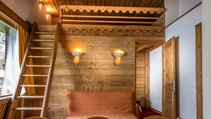 Un intérieur en bois comprenant un lit, une table, des chaises, un escalier et deux lampes fixées au mur.