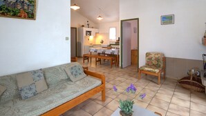 A cozy living room with a sofa, a dining table, and chairs. There is a kitchen in the background with a refrigerator and a sink.