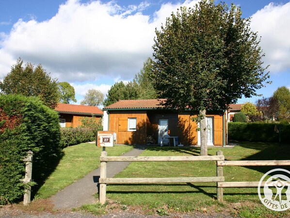 A wooden cabin with a red roof, surrounded by greenery and a wooden fence.