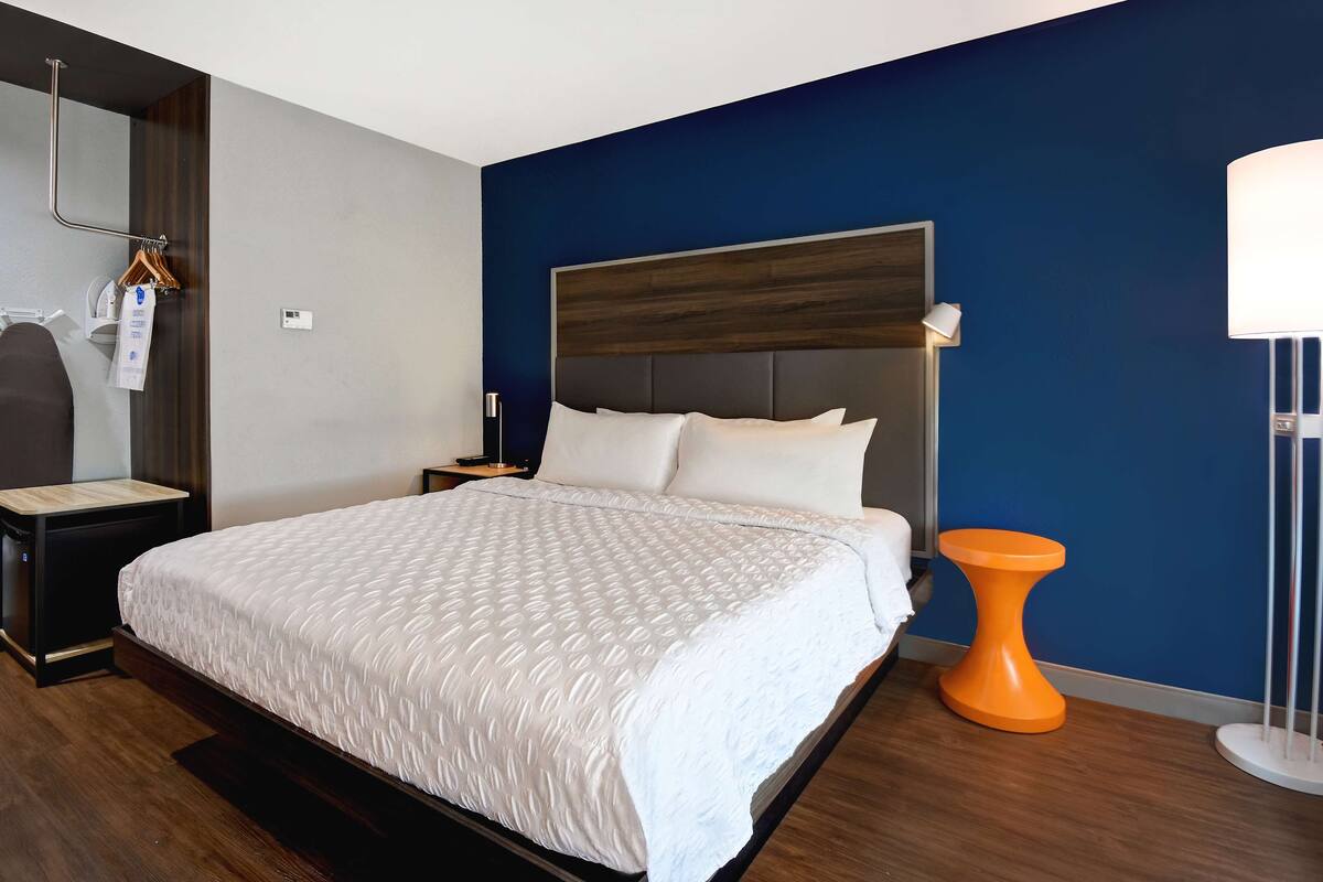 Une chambre d’hôtel moderne avec un grand lit, une table de chevet, un miroir, une armoire et une petite table d’appoint de couleur orange vif.