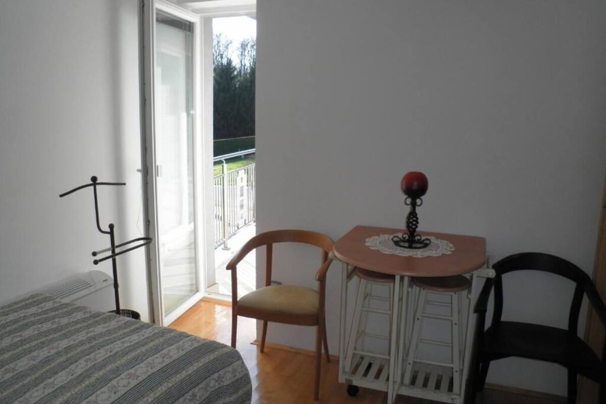 A small room with a bed, a chair, a table with a lamp, and a sliding glass door leading to a balcony.