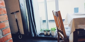 A room with a brick wall, a scooter, a window with curtains, a wooden chair, a table with a plate and a cup, and potted plants on the windowsill. - MalinaApt SuperLoft (Kyiv)