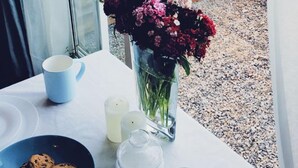 Una mesa con platos, una taza azul, una taza blanca, un vaso de leche, un plato azul con galletas y un florero con flores.