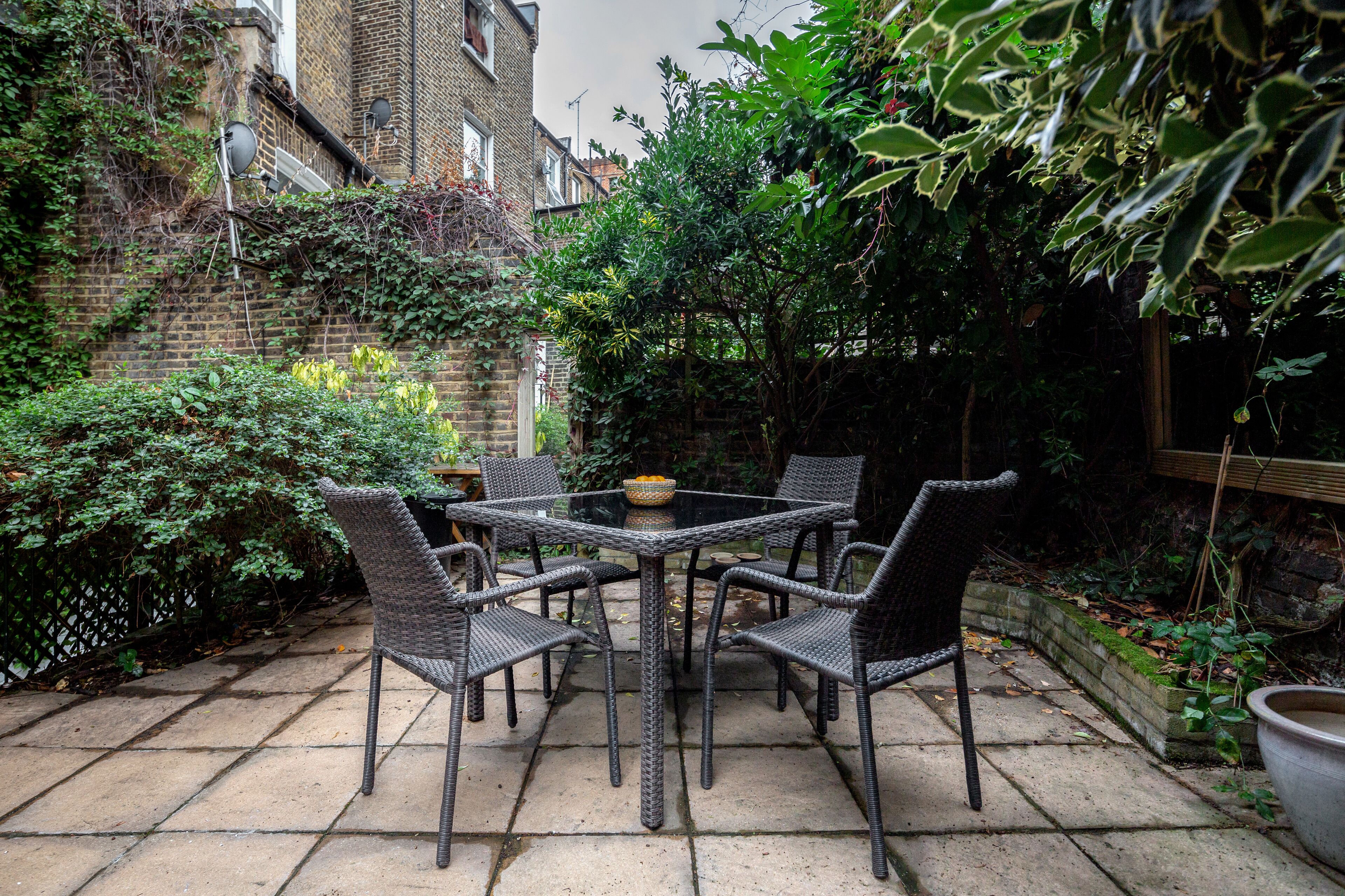 A patio with a glass table surrounded by wicker chairs, set against a brick wall with greenery and a wooden fence.