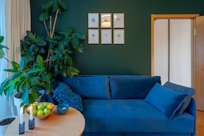 A room with a blue sofa, a round wooden table with a bowl of fruit and candles, a large plant, and framed pictures on the wall.