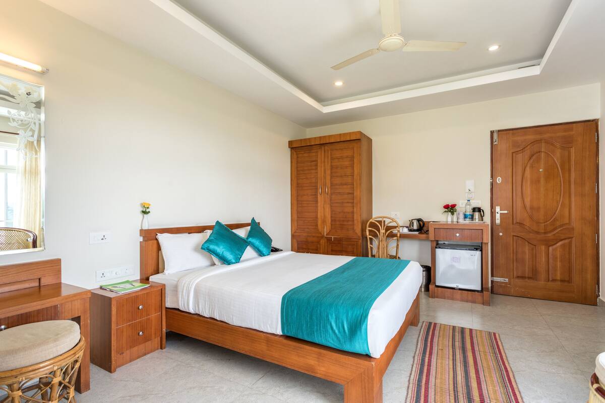 A bedroom with a wooden bed, a desk, a chair, a wardrobe, a small refrigerator, and a ceiling fan.