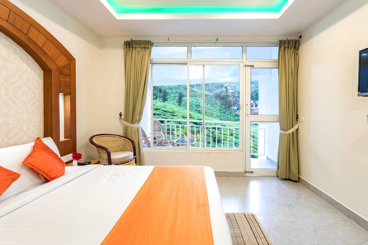 A hotel room with a large bed, a wooden headboard, a balcony with a view of green hills, and a television on the wall.