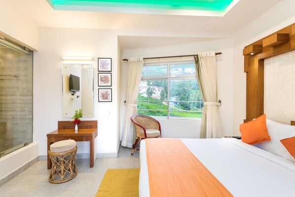A bedroom with a bed, a desk, a chair, a TV, and a window with curtains. - Monsoon Grande (Devikolam)