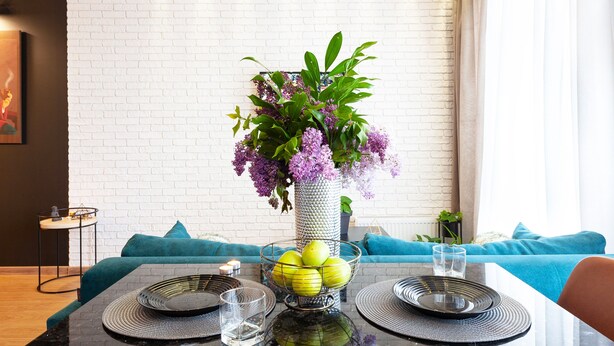 Een eetgedeelte met een witte bakstenen muur, een blauwe bank, een tafel met een bloevas en een fruitschaal.