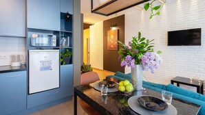 Een moderne keuken met een ingebouwde magnetron, koelkast en planken. Er is een eethoek met een tafel, stoelen en een vaas met bloemen. Een televisie is aan de muur bevestigd.
