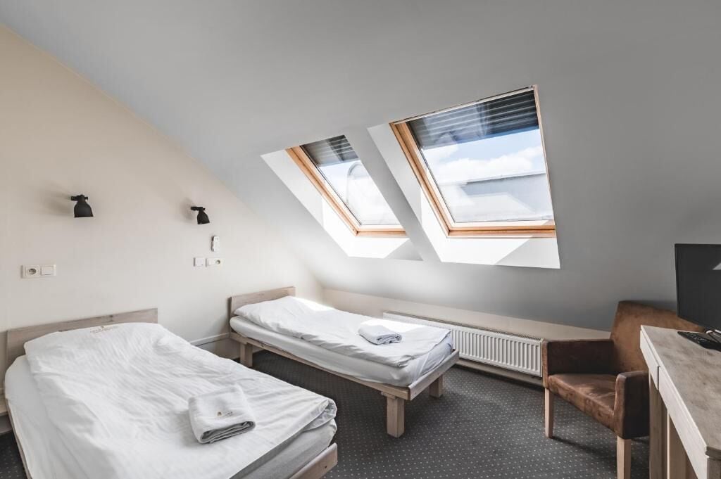 Una habitación pequeña y moderna con dos camas individuales, un escritorio y una silla. Cuenta con tragaluces que proporcionan luz natural.
