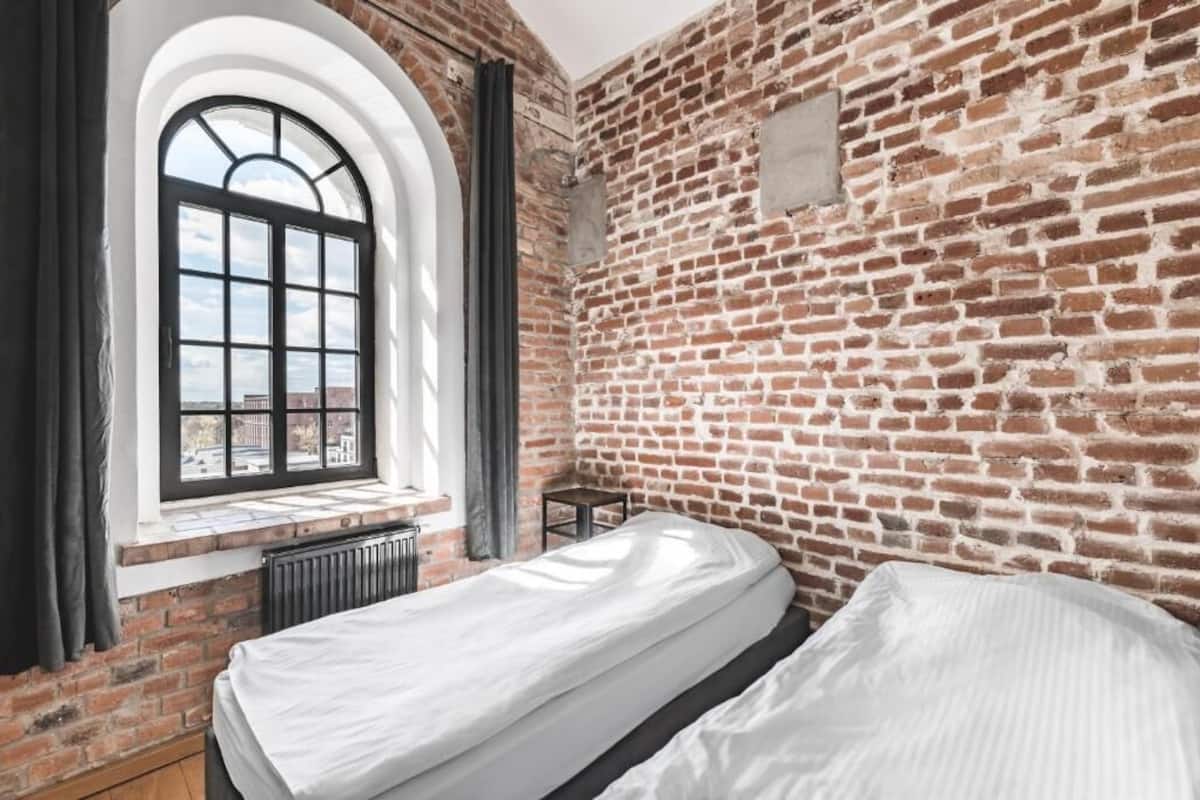 Una habitación con pared de ladrillo, ventana con vistas, cama y una mesita.