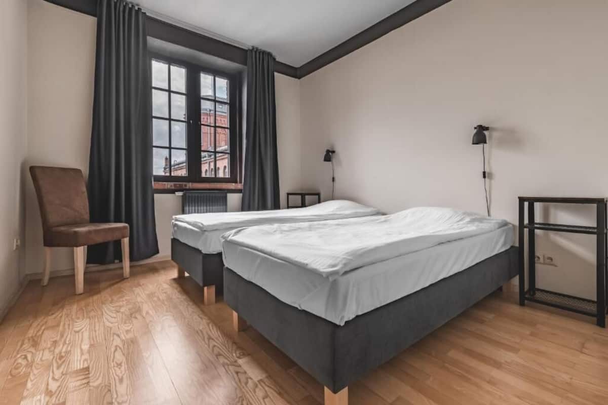 Una habitación con dos camas, una silla, un ventanal con cortinas y un radiador.