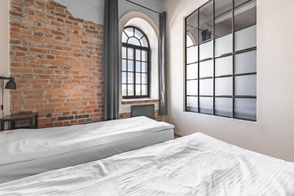 Una habitación con dos camas, una pared de ladrillo, un ventanal grande y un radiador.