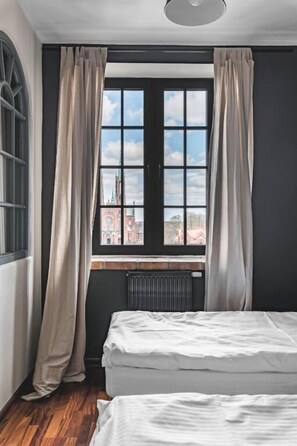 A room with two beds, a window with curtains, a radiator, and a view of a building outside. - Aparthotel Stara Przędzalnia (Żyrardów)