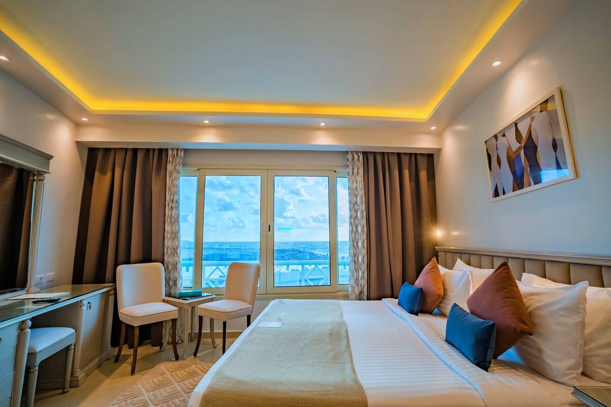 Une chambre d’hôtel équipée d’un lit, d’un bureau, de chaises et d’une grande fenêtre donnant sur un paysage agréable.