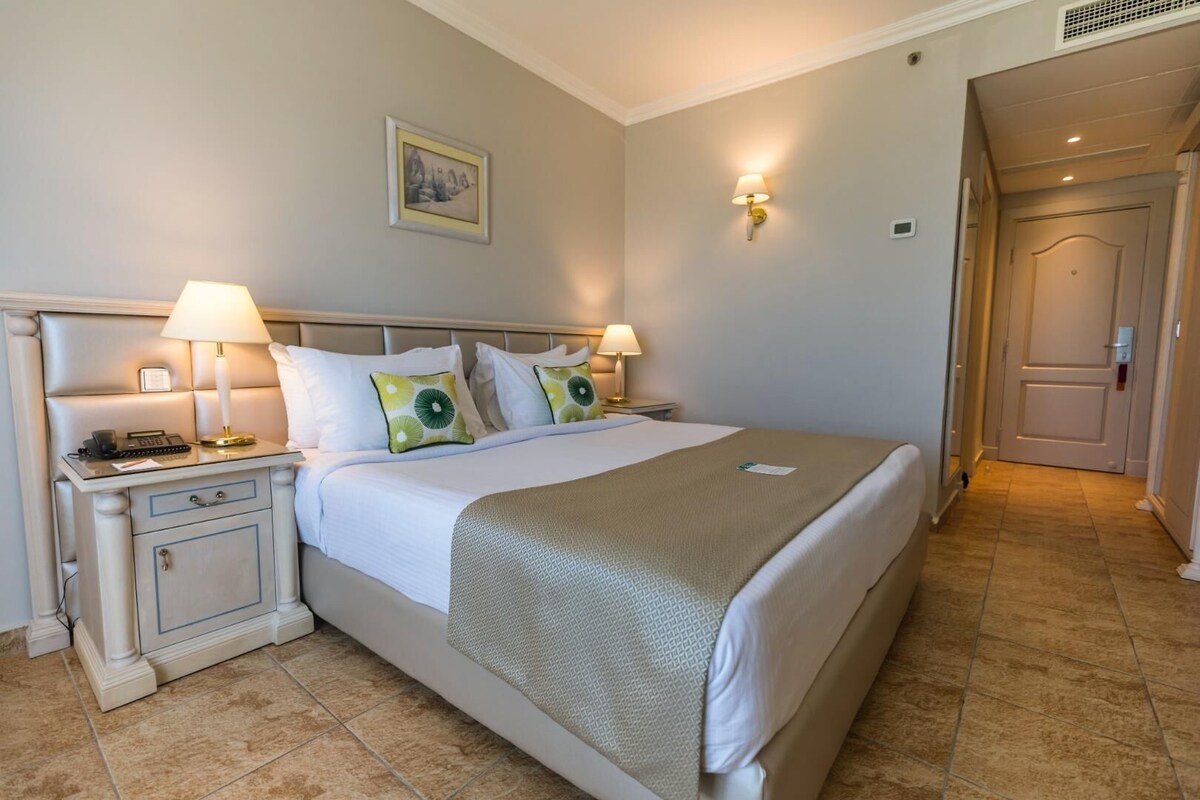 A hotel room with a bed, bedside tables, lamps, a painting on the wall, and a door.