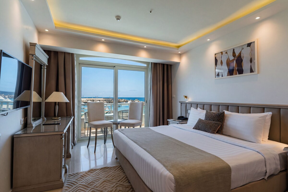 A hotel room with a bed, a desk with a lamp, a TV, a large window with curtains, and a view of the sea.