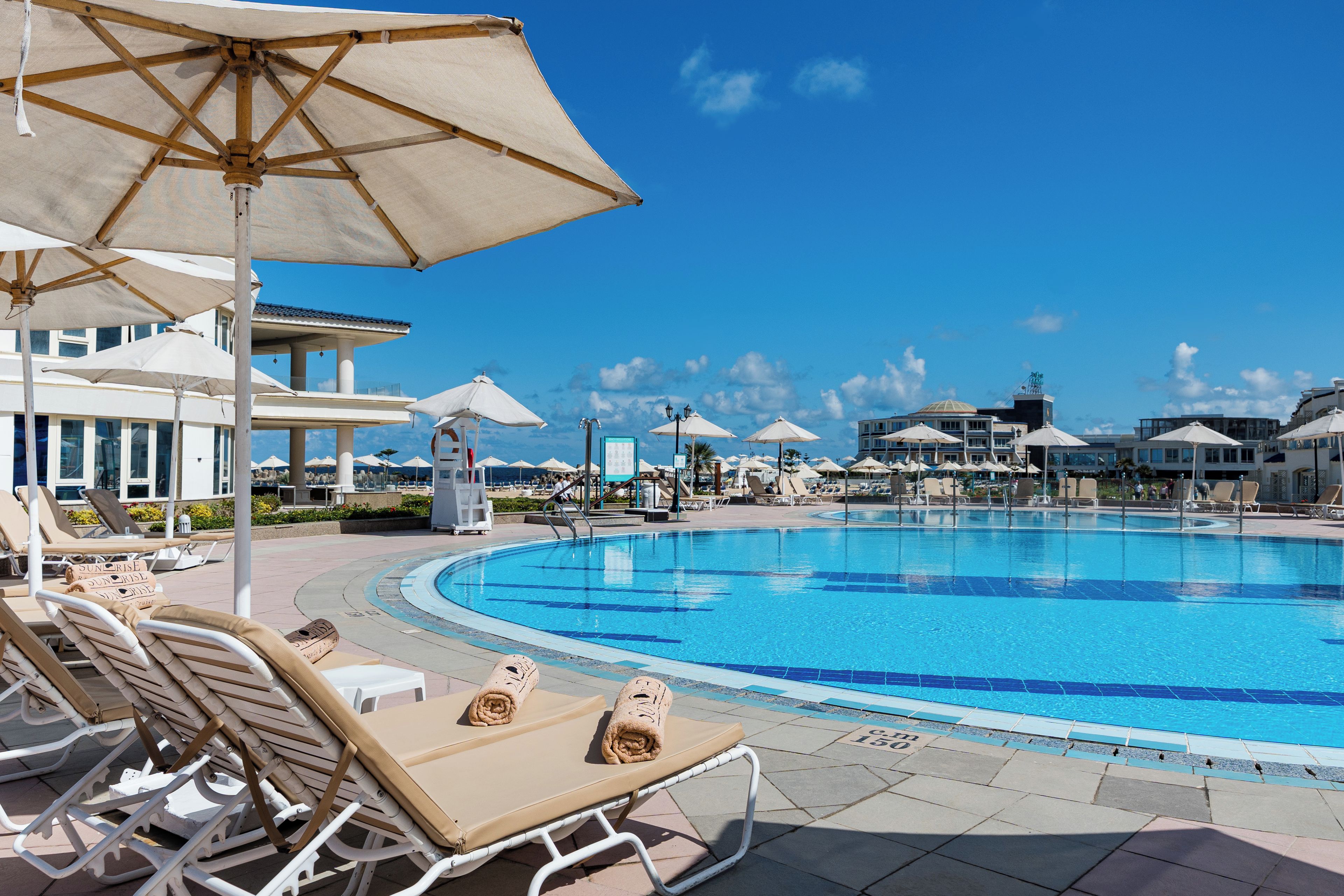 A swimming pool with lounge chairs and umbrellas, surrounded by buildings and a clear sky.