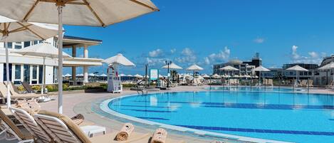 A swimming pool with lounge chairs and umbrellas, surrounded by buildings and a clear sky.