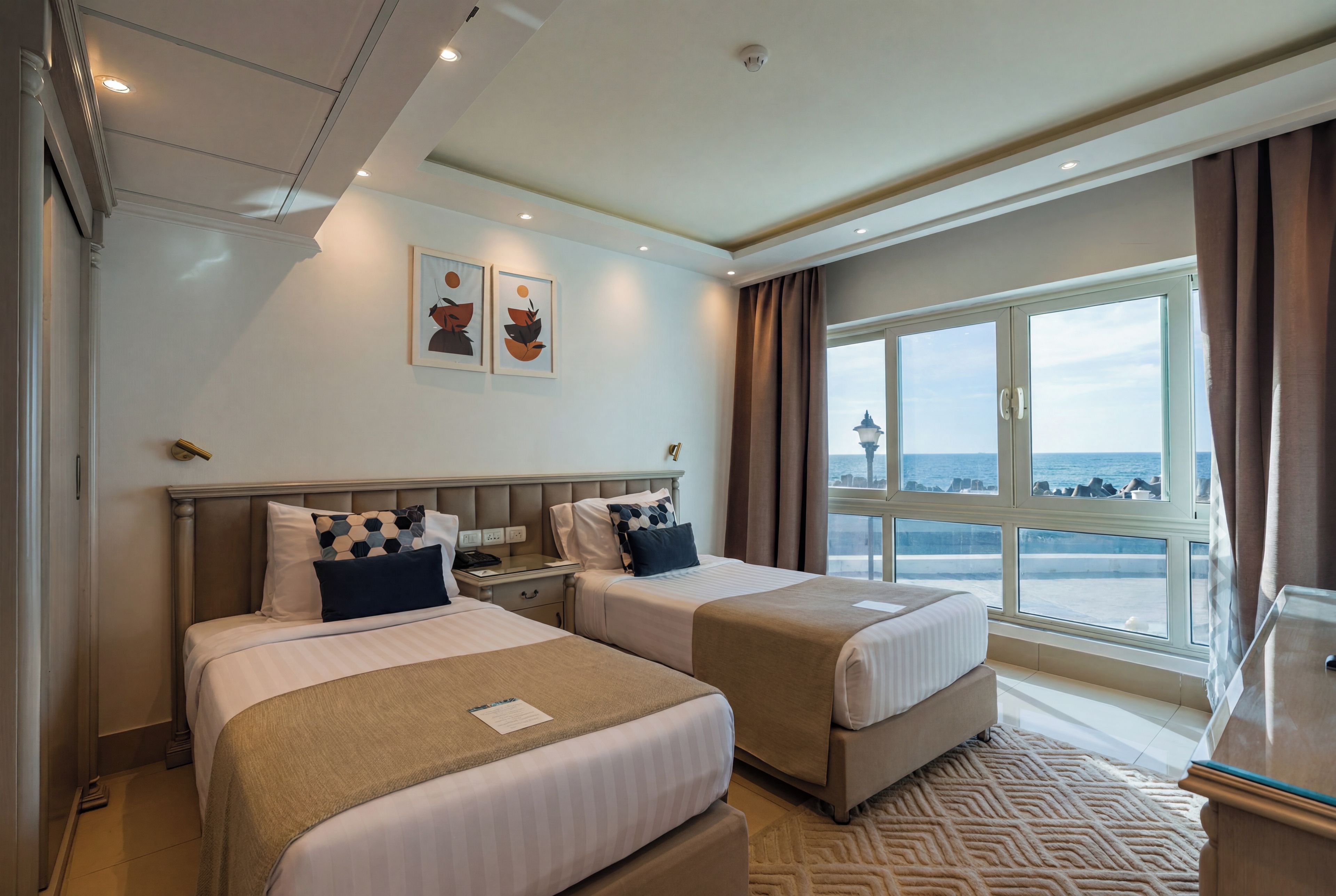 Habitación de hotel con dos camas, un ventanal con vista al mar e iluminación empotrada en el techo.
