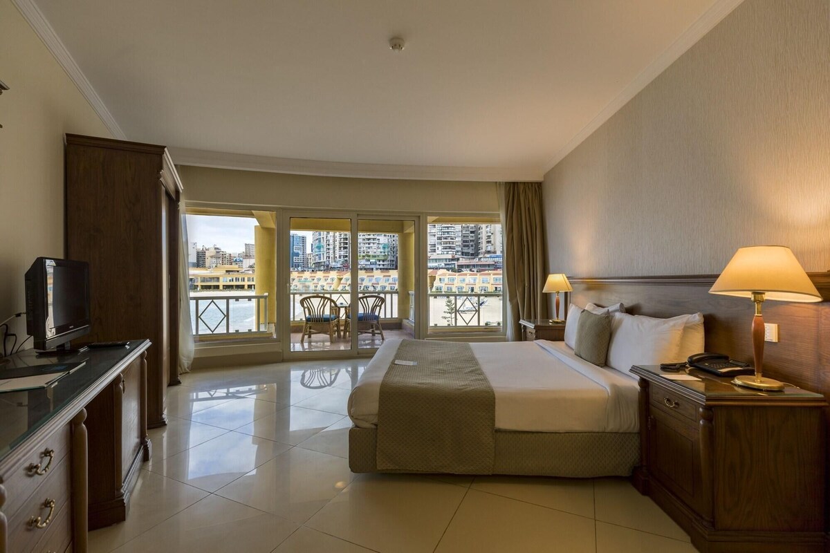 A hotel room with a bed, bedside tables, a TV, a balcony with chairs, and a city view.
