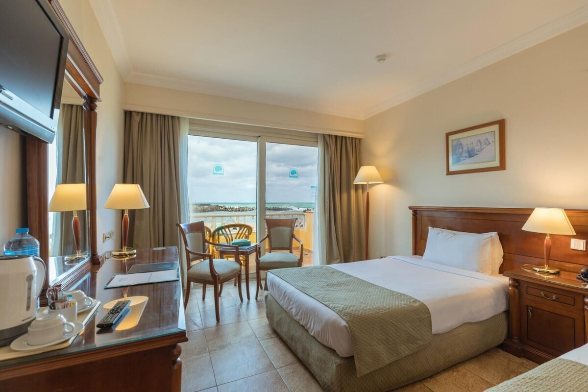 A hotel room with a bed, a desk with a computer, a chair, and a view of the beach.