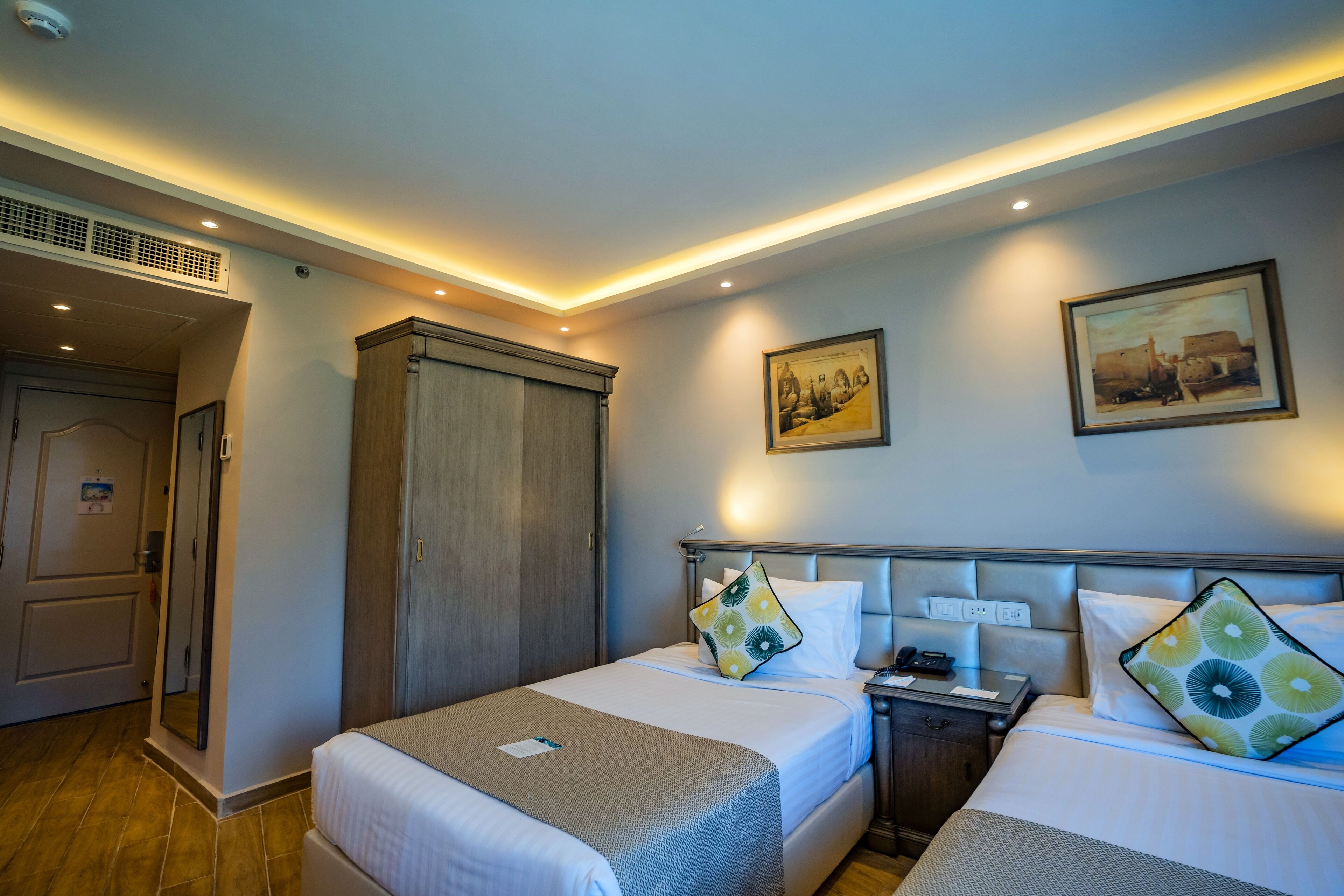 Quarto de hotel com duas camas, um guarda-roupa, uma mesinha de cabeceira e um quadro na parede.