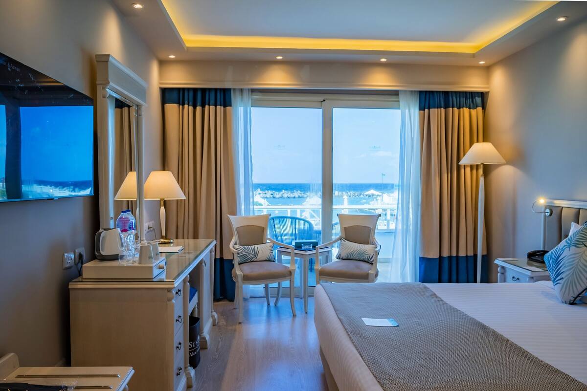 Une chambre d’hôtel avec un lit, deux chaises, un bureau avec une lampe, une télévision et une vue sur la mer.