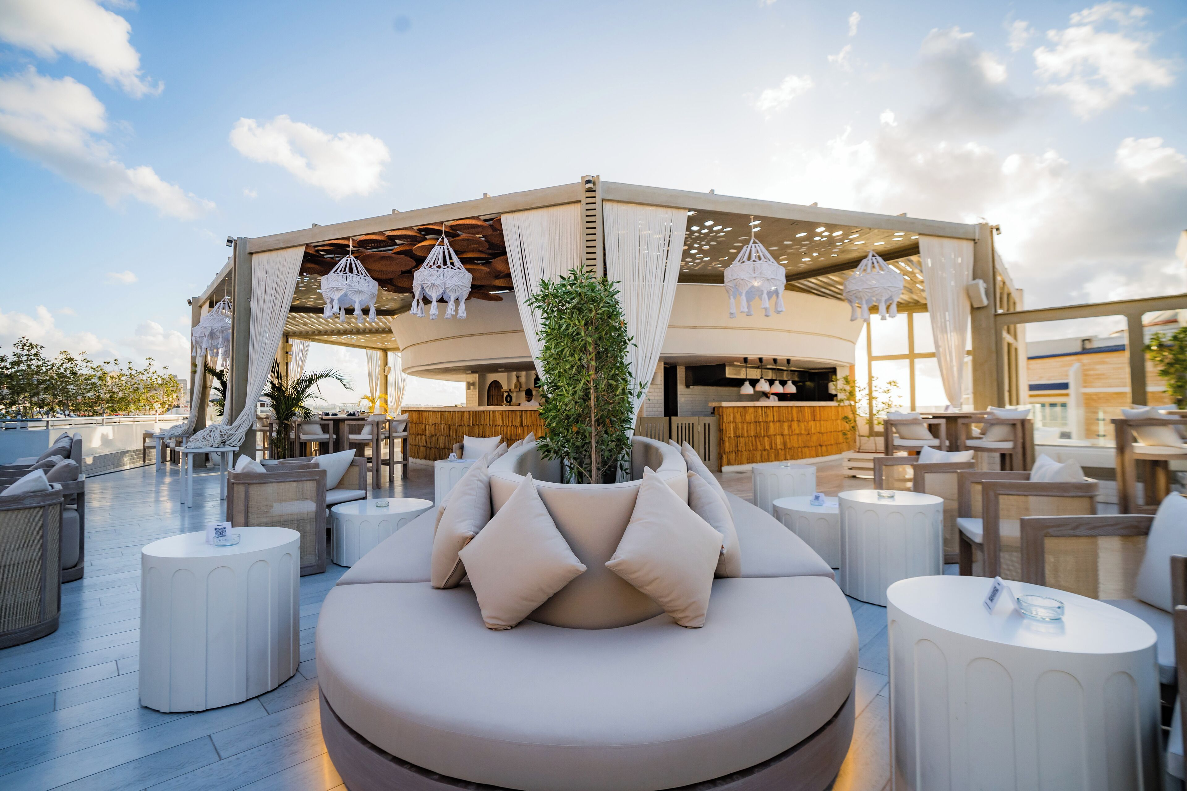 A rooftop lounge area with round seating, a central planter, and a bar in the background.
