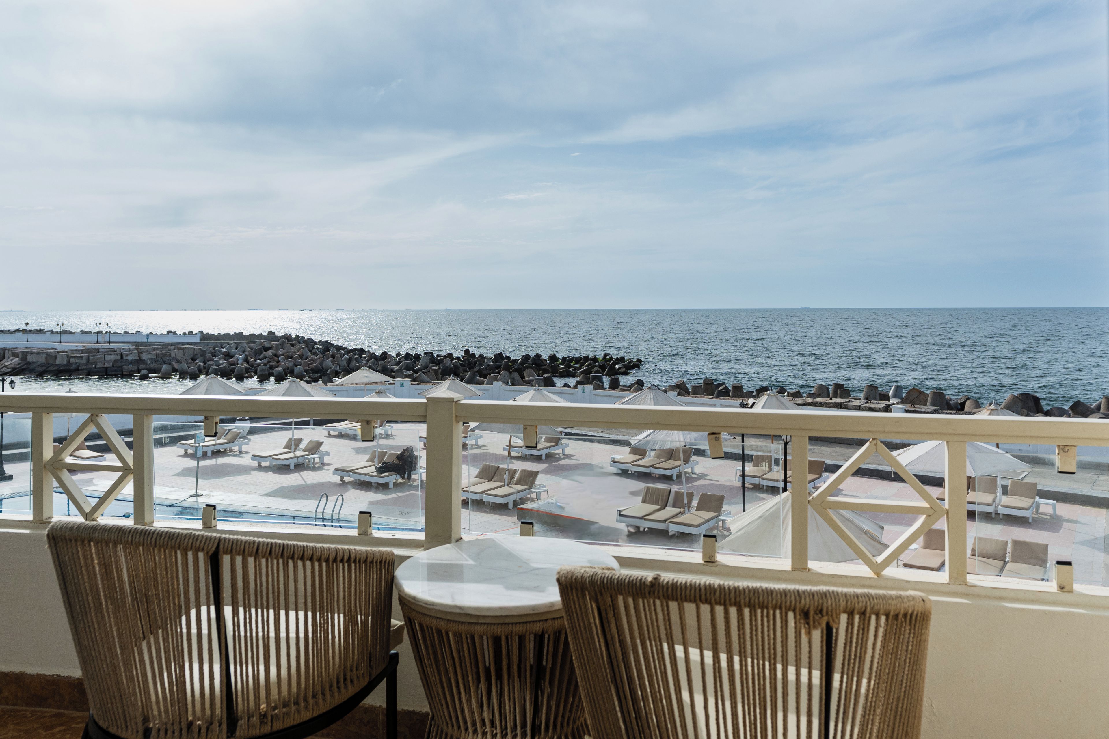 En balkon med fletstole og et bord, med udsigt over en pool og havet.