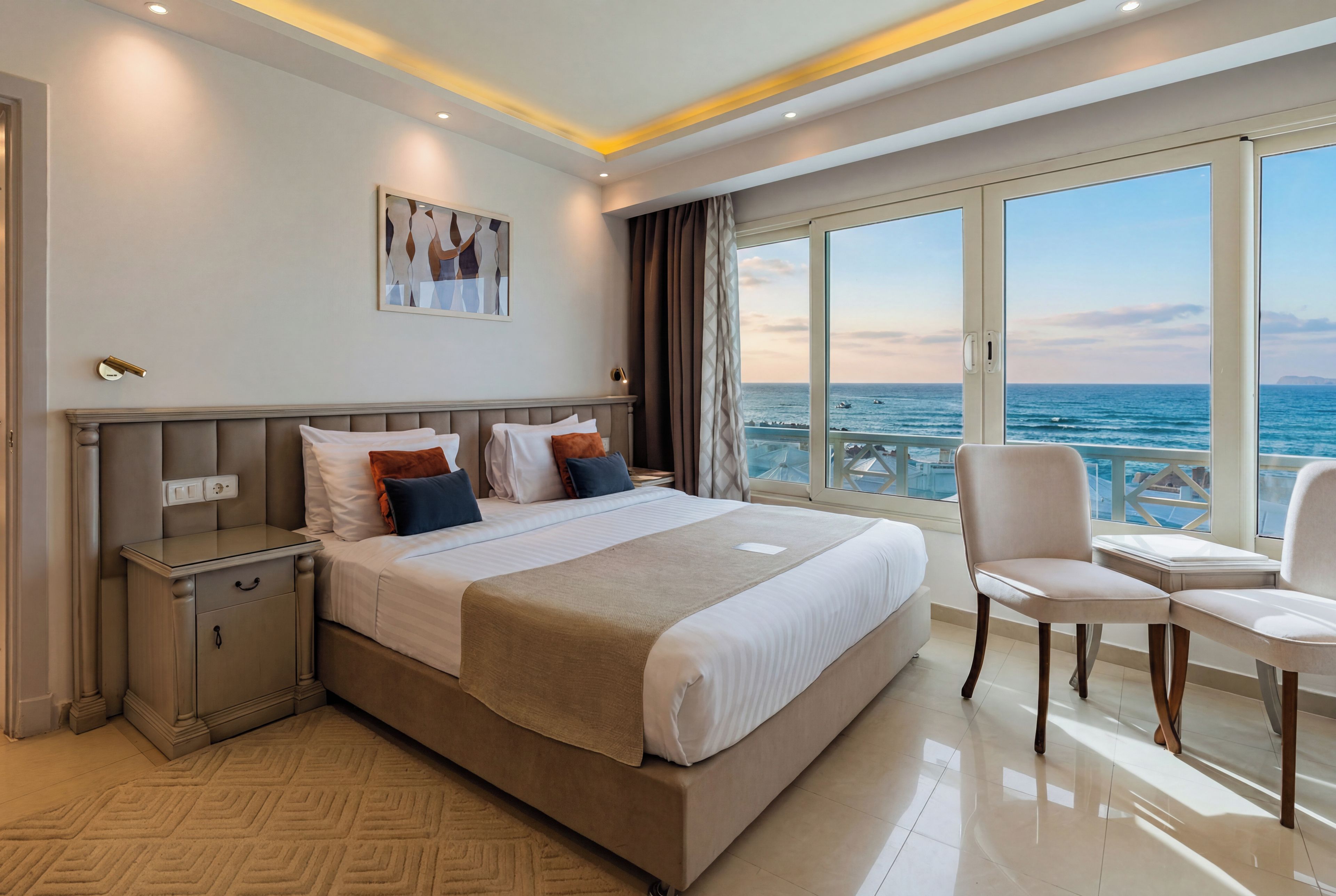 A hotel room with a large bed, a bedside table, a chair, and a view of the sea.