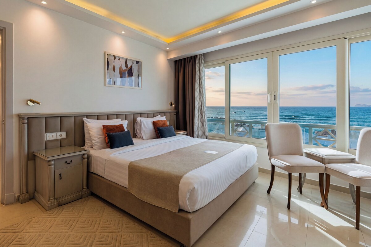 A hotel room with a large bed, a bedside table, a chair, and a view of the sea.