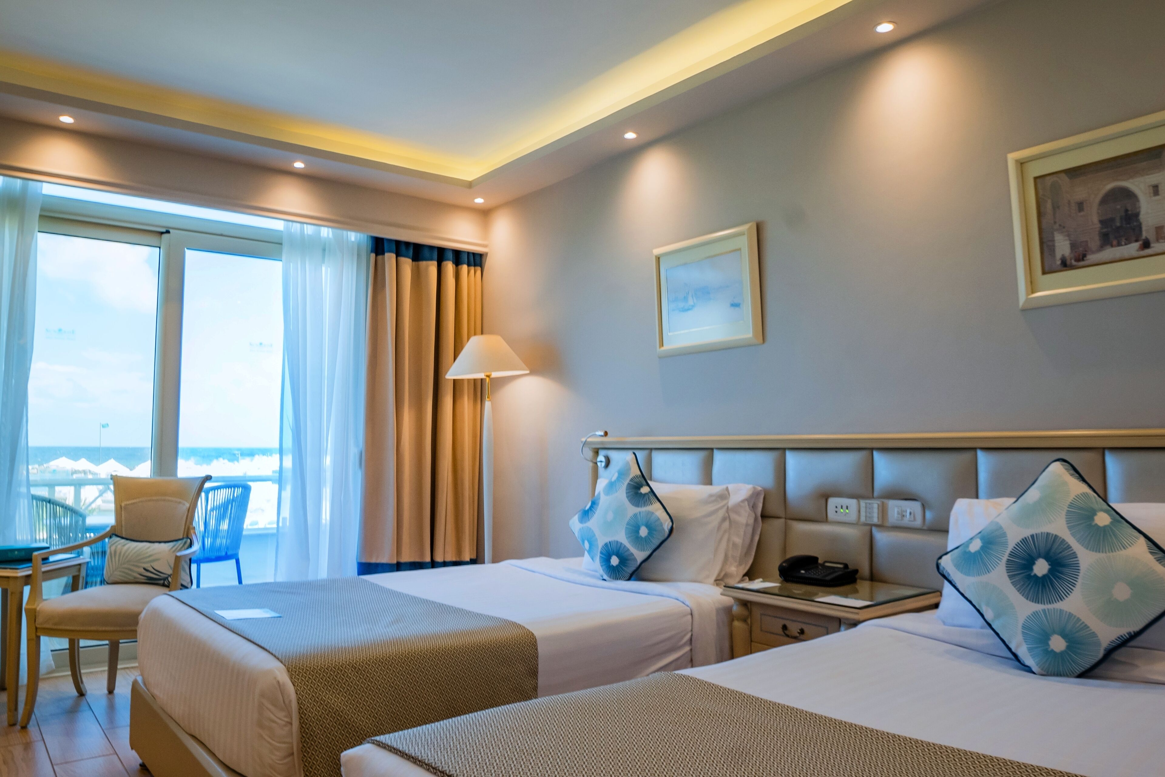 A hotel room with two beds, a desk, and a chair. There is a window with curtains, a lamp, and a painting on the wall.