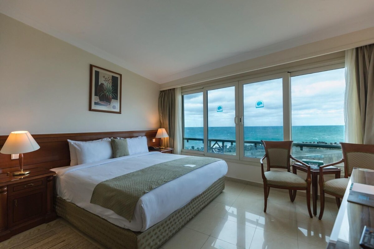 A hotel room with a large bed, two bedside lamps, a chair, a desk, and a view of the ocean.