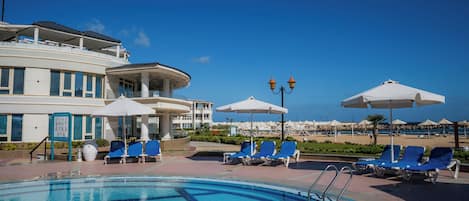 A swimming pool with blue water, surrounded by lounge chairs and umbrellas, with a building in the background.