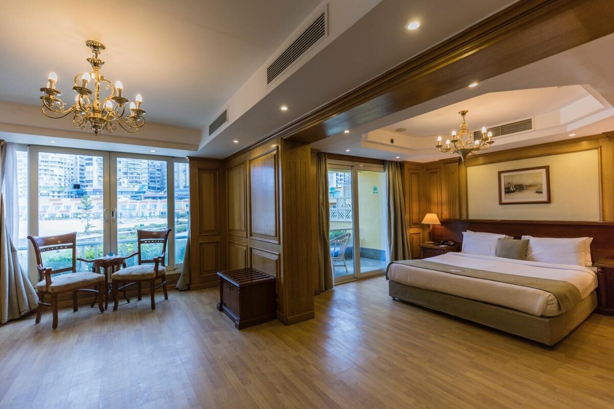 A hotel room with a large bed, a desk with chairs, a chandelier, and a view through the window.