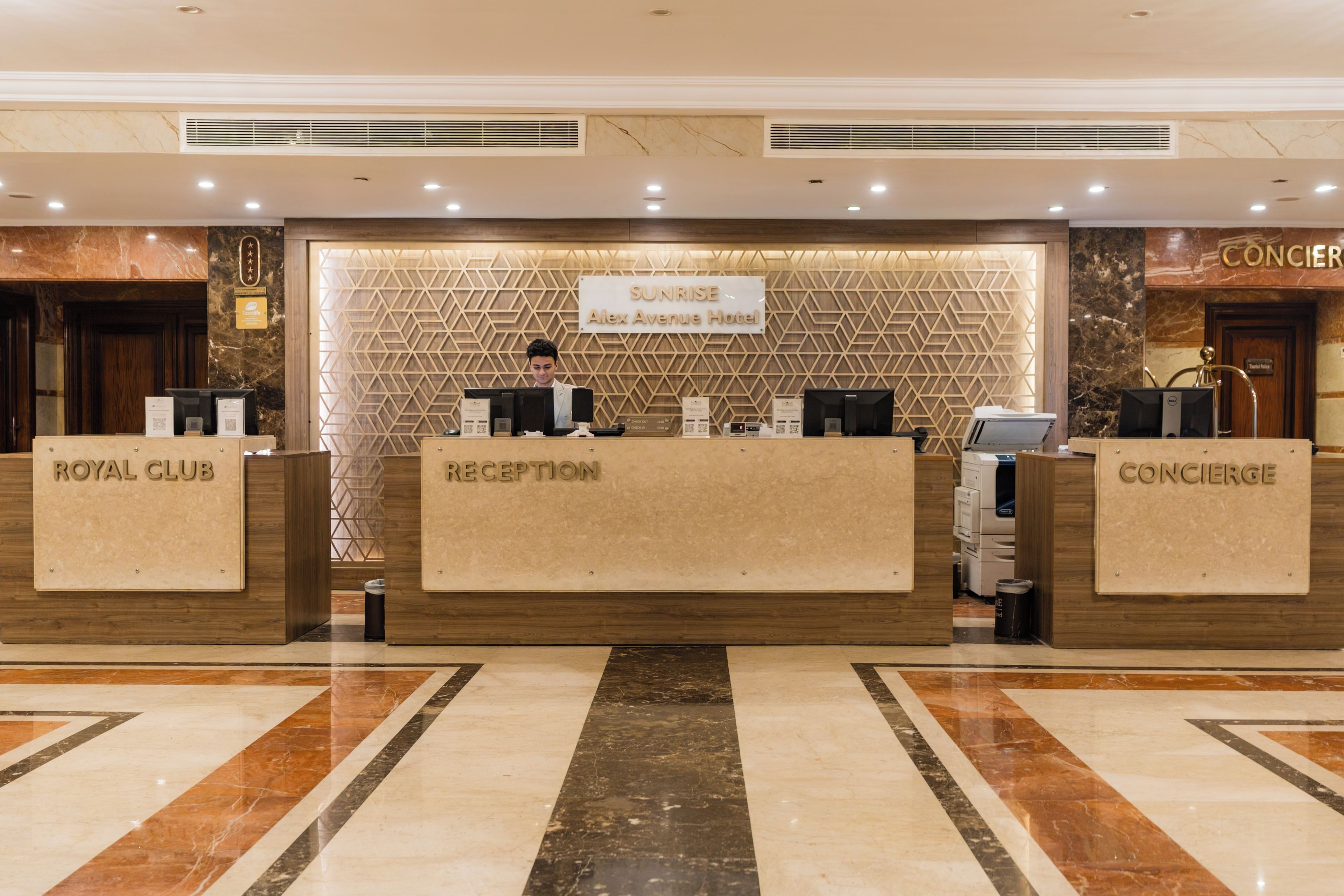 A hotel reception area with a reception desk labeled 'Sunrise Alex Avenue Hotel,' flanked by 'Royal Club' and 'Concierge' counters.