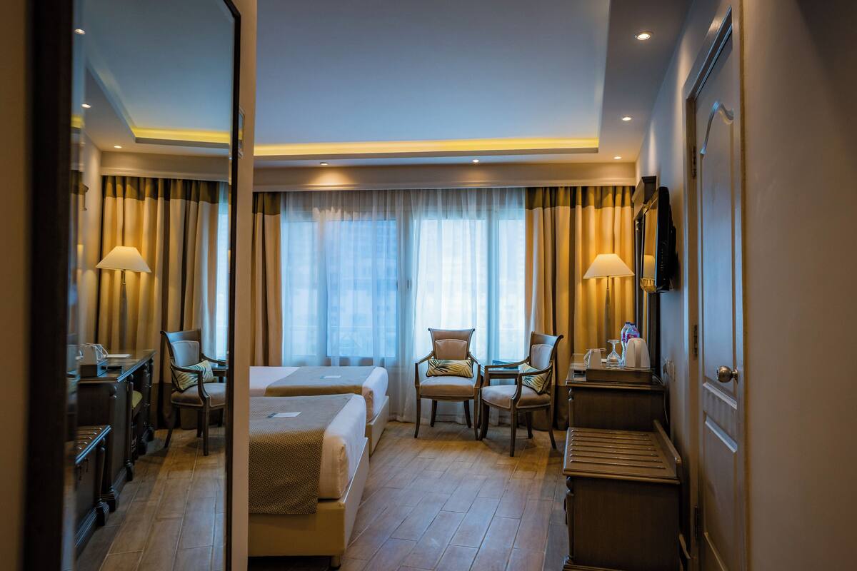 Une chambre d’hôtel avec deux lits, un bureau, des chaises, une télévision et une grande fenêtre avec des rideaux.