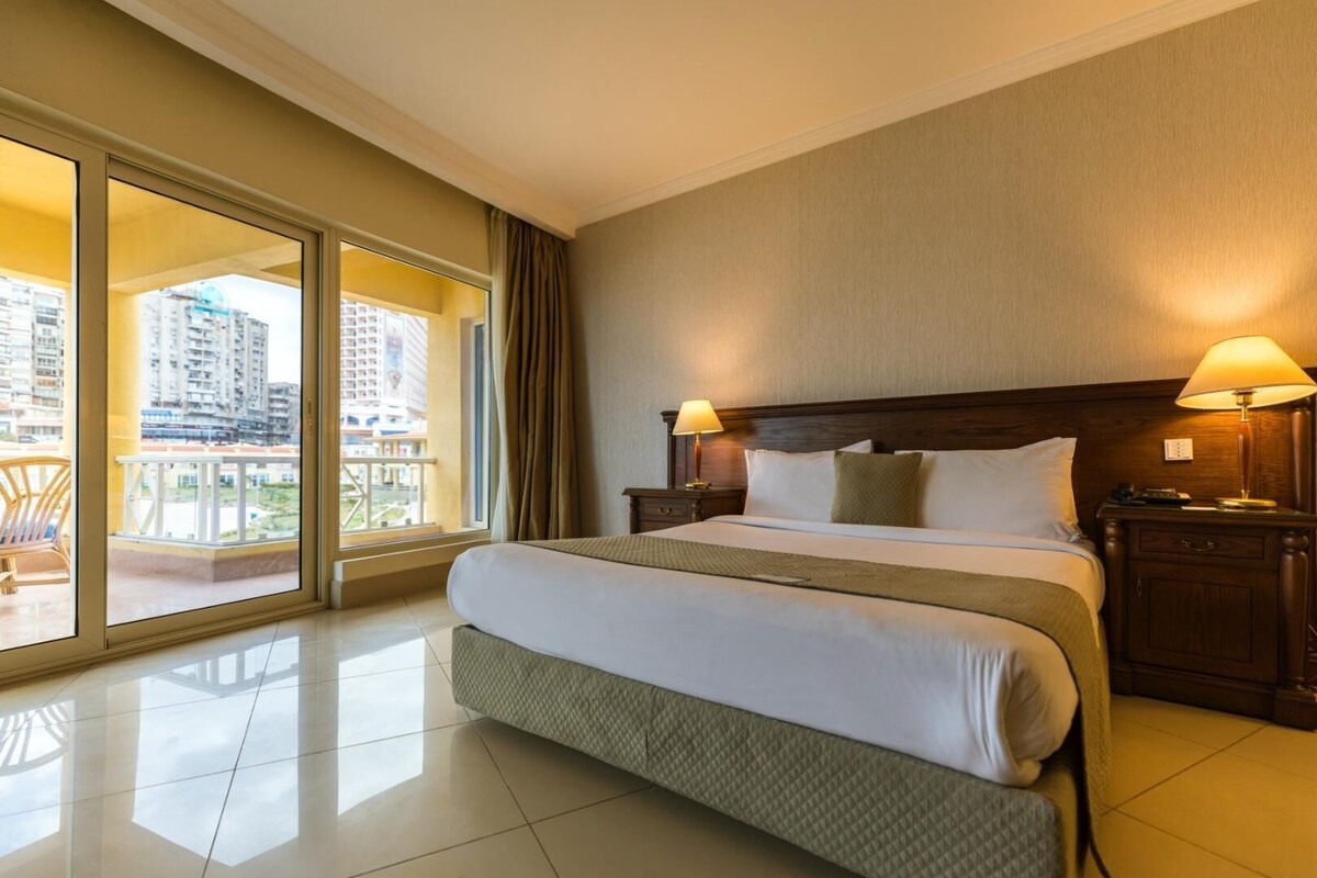A hotel room with a large bed, two bedside lamps, a wooden nightstand, and a view of high-rise buildings through a sliding glass door.
