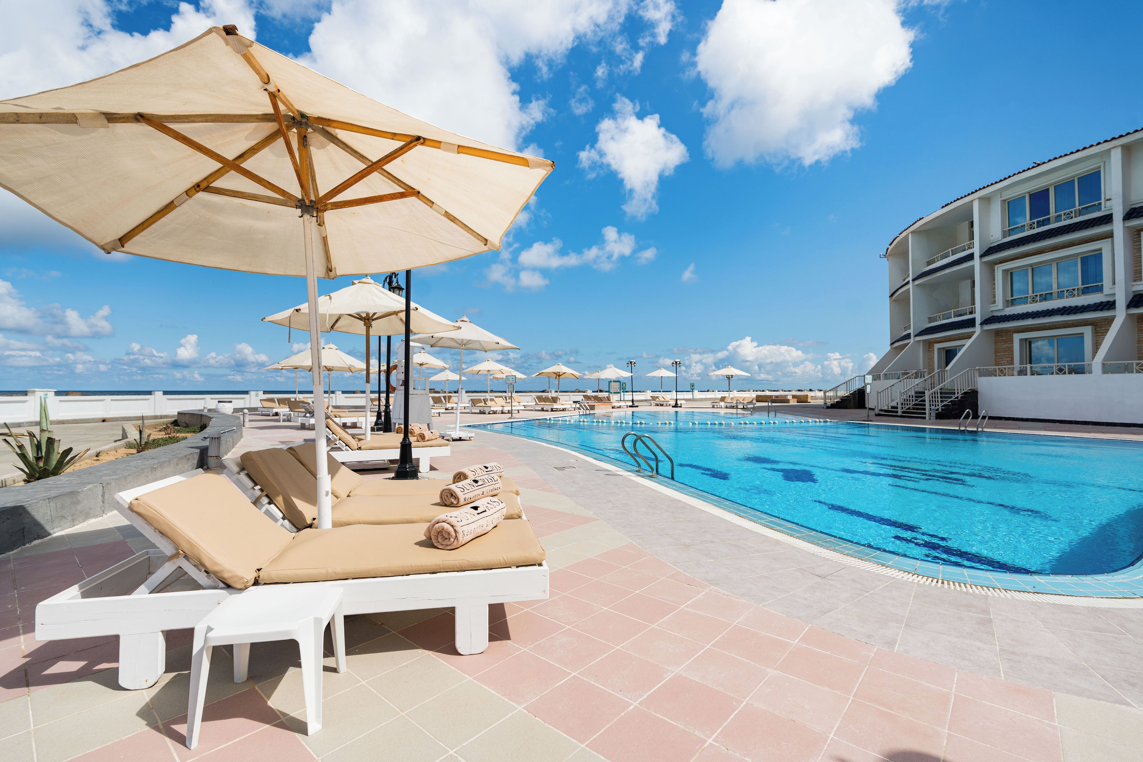 A poolside area with lounge chairs and umbrellas, a swimming pool, and a building with multiple balconies.