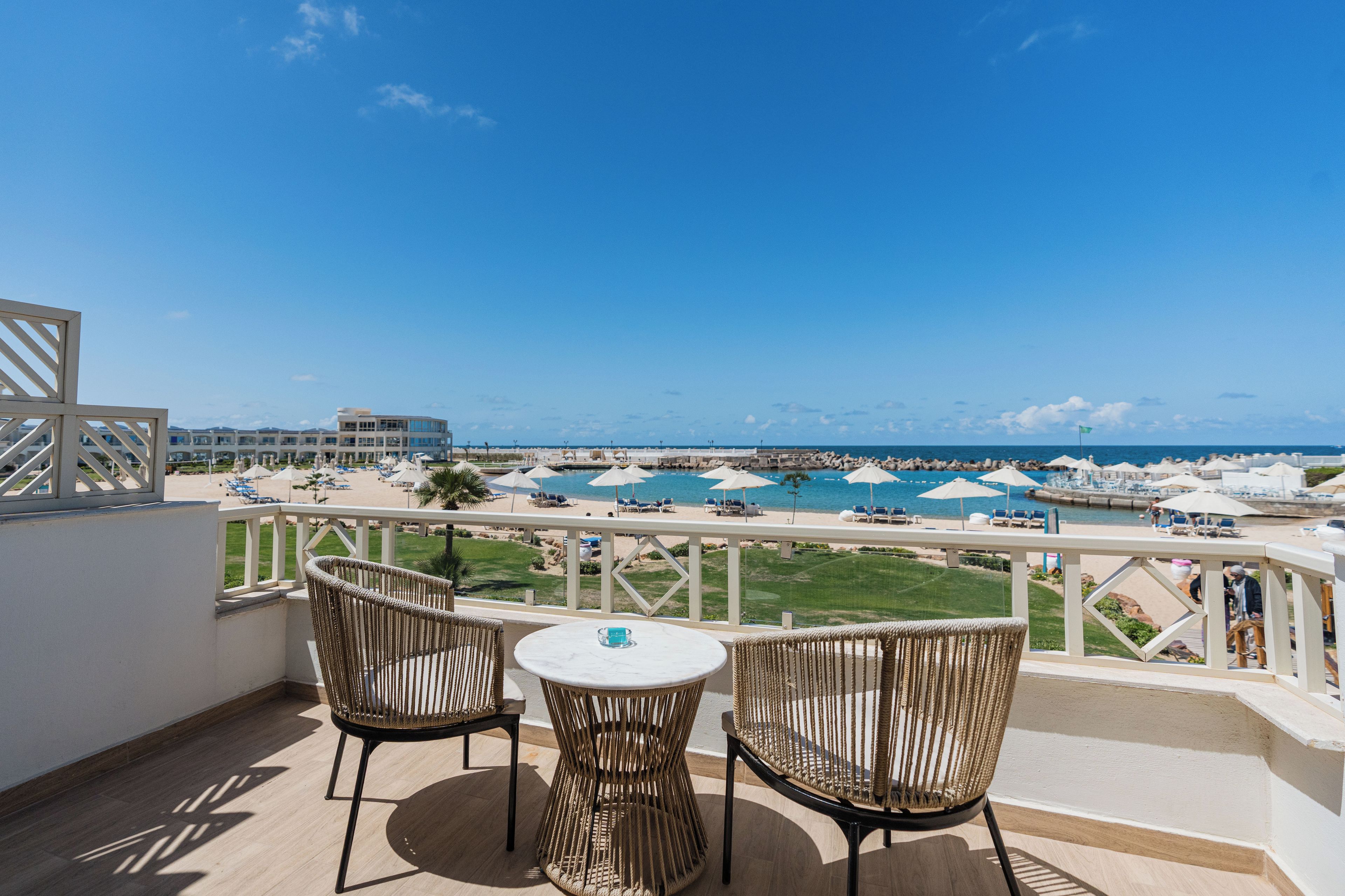 A balcony with two wicker chairs and a small table, overlooking a beach with umbrellas and lounge chairs.