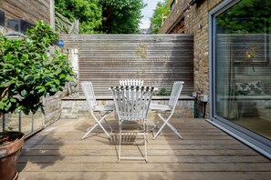 Un patio con una terraza de madera, una mesa de metal blanco y dos sillas de metal blanco.